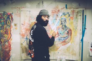 A young male artist with a beard, wearing a beanie and glasses in front of a graffiti style painting of a human figure.