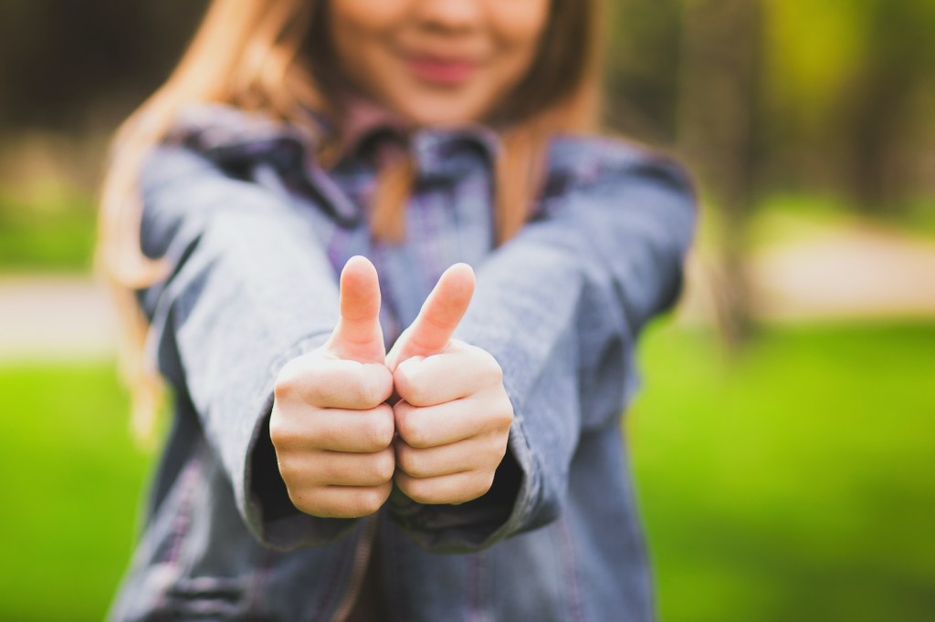 Young teenage girl giving thumb up as sign of success. Portrait of happy smiling anonymous caucasian child dressed in blue jeans casual clothing. Focus at hands.