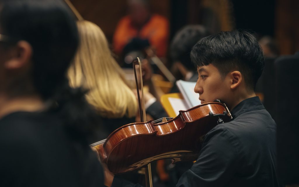 Student musician from the University of Melbourne Symphony Orchestra. Photo of a young person with black hair playing a violin among other musicians.