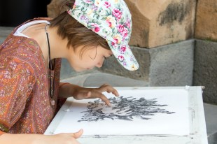 Artist. A young girl in floral baseball cap leaning over charcoal drawing using her finger to draw. Child artist taking a risk.