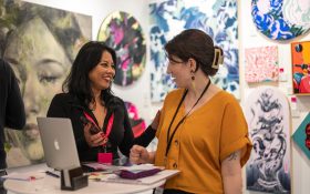 Affordable Art Fair. Image is two women smiling and talking to each other about art works, with art on the walls behind them. One woman is wearing a mustard colour short sleeved shirt and has brown hair held back in a clip and a lanyard, the other has longer dark hair and a black shirt with red lanyard.