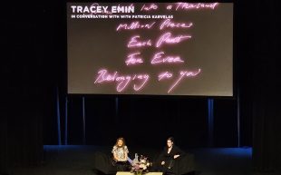 Live and in Conversation After-Hours: Tracey Emin hosted by the NGV leading up to the 2023 NGV Triennial. Photo: ArtsHub. Two figures sitting on the stage in a large auditorium. A projector is showing the words ‘Tracey Emin’ against pink neon phrases that form an artwork.