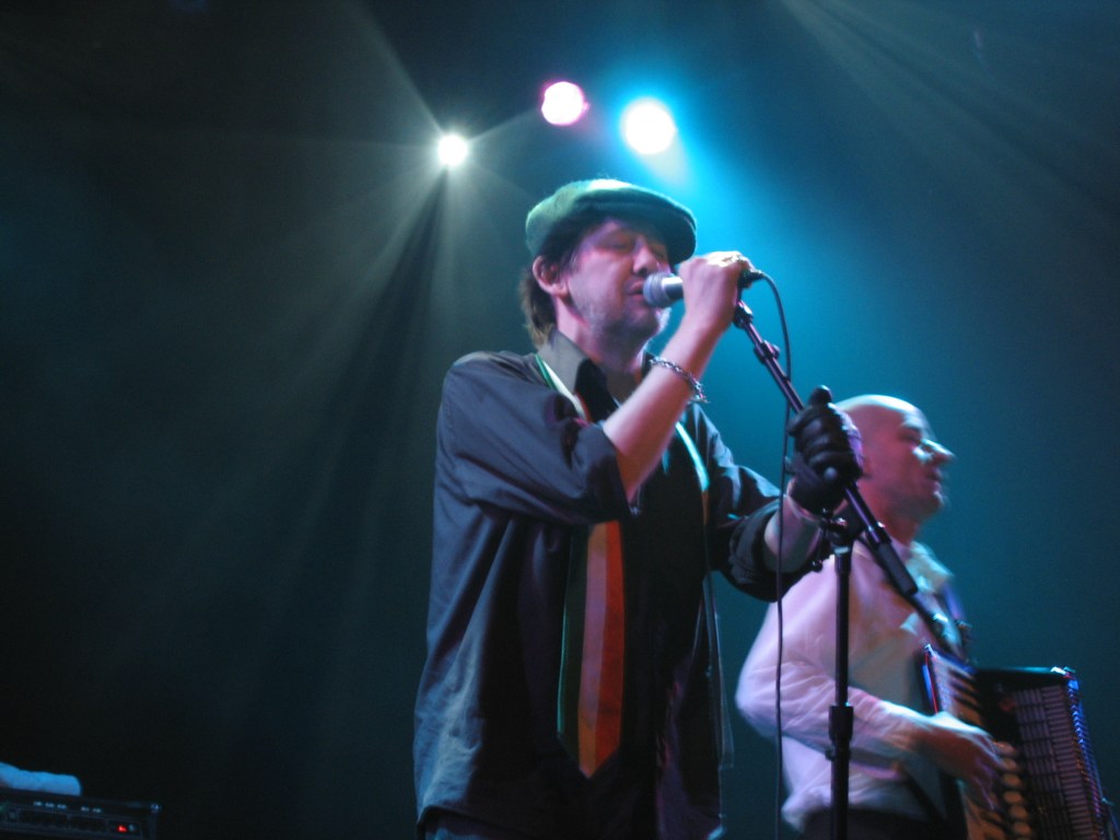 Shane MacGowan. Image is of two band members on a stage, one singing into a microphone and wearing a flat cap.