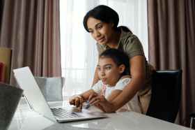 AI. Woman with brown skin leans over her daughter to teach AI on laptop.