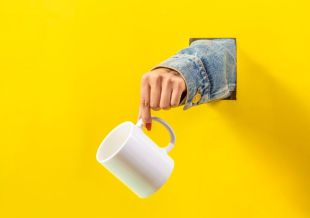 an arm coming through a bright yellow wall, hand holding a white mug.