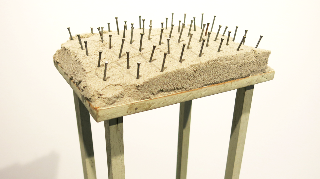 Alison McKay, Untitled, 2017, Sand and nails on wooden stand. Image: Supplied. An artwork consisting of a tiny precarious plinth with nails hammered into the top, and loose damp sand surrounding the nails.