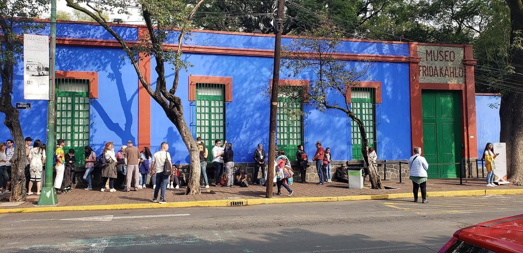 Frida Khalo Museum exterior. Image: Wikimedia Commons. A blue rectangular building shown from street view where people are gathered outside.
