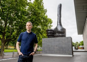 Man in blue t-shirt standing next to outdoor bronze sculpture of a huge hand with an elongated thumb sticking up in the air. David Shrigley, NGV