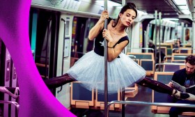 A young woman in a tutu is in an underground railway carriage and swirling around a pole.