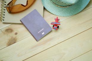 overseas literary residencies. Image is of a passport, a little toy plan, a sunhat, notebook and sunglasses - all on the wooden boards of a tabletop.