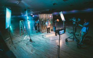 entry-level arts jobs. Photo: Jakob Owens, Unsplash. A figure stands in the centre of a dark indoor space with a photography set-up. Lets are shining on him and people are sorting out technical things with a camera.