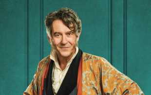 A man with a white shirt, black jacket and an orange dressing gown against a turquoise backdrop. Adrian Lukis in Being Mr Wickham.