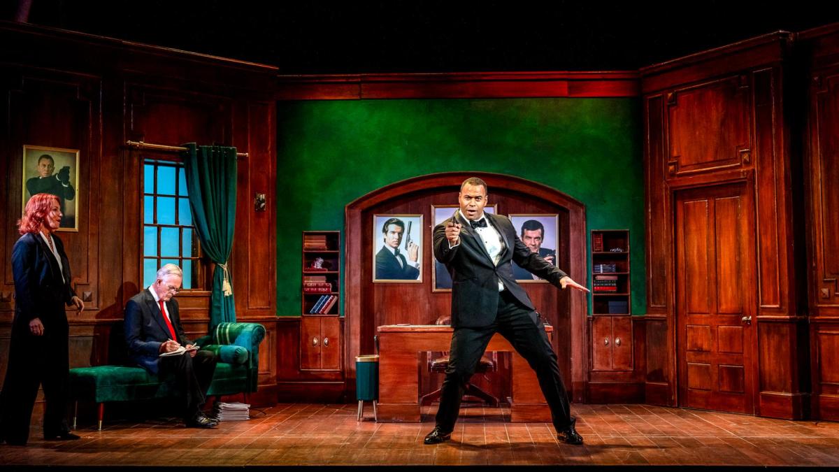 A black man in a tux holding a gun in a drawing room. There's also another man and a woman there in this production of A Role to Die for.