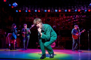 A scene from 'Sunny Afternoon', the musical about and featuring the music of The Kinks at The Alexandra, Birmingham. A green-glad singer crouches centrestage beneath the spotlight and sings into a microphone; three other musicians, dressed similarly, can be seen behind him.