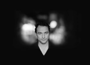 Promotoinal image for 'Playing Burton' with actor Matthew Rhys. The moody black & white photo shows Rhys' sharp-cheekboned fair-skinned face emerging from the darkness. He has tousled black hair and wears a black shirt.