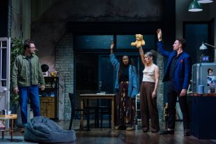 A scene from 2:22 – A Ghost Story at the Theatre Royal Bath. Three characters stand with their hands raised, one holding a teddy air, and face a fourth character in an open plan stage kitchen.